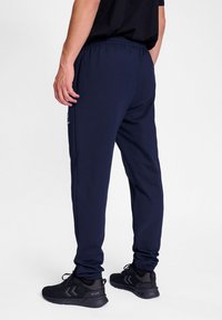 Marineblaue Baumwoll-Jogginghosen mit elastischem Taillenbund und Bündchen an den Knöcheln, mit Gesäßtasche und dezentem Logodetail. Getragen mit schwarzen Sportschuhen.