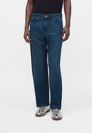 Homme portant un jean droit bleu foncé et des baskets argentées métalliques, debout devant un fond blanc.
