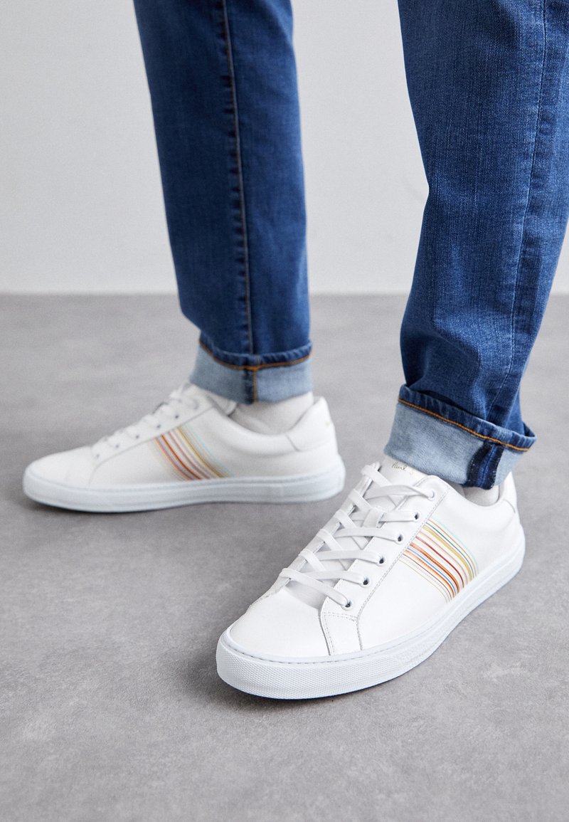 Baskets en cuir blanc avec des rayures multicolores, design à lacets, semelle en caoutchouc et col rembourré, associées à un jean en denim bleu.