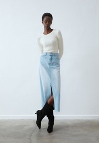 Jupe en denim bleu clair avec une fente avant, associée à un haut ajusté à manches longues en coton ribbed blanc et des botteurs noirs à bout pointu.