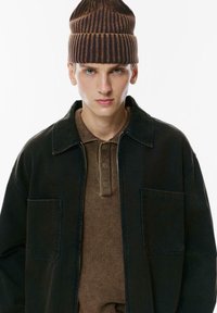 Jachetă din denim negru cu două buzunare frontale peste un tricou polo texturat maro cu trei butoane, completată cu o căciulă din lână cu dungi maro.