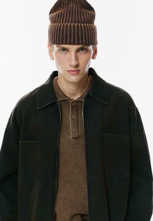 Black denim jacket with two front pockets over a brown textured polo shirt featuring three buttons, topped with a brown striped beanie.