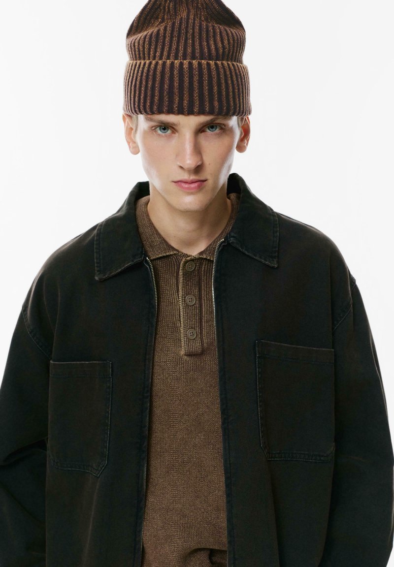 Jachetă din denim negru cu două buzunare frontale peste un tricou polo texturat maro cu trei butoane, completată cu o căciulă din lână cu dungi maro.