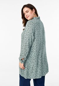 Long-sleeve shirt in light green fabric with a floral pattern. Features a collar and is loose-fitting with rolled cuffs.