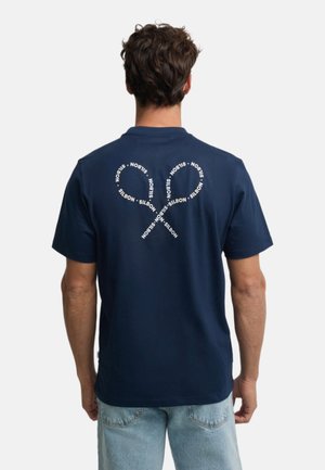 Hombre con camiseta azul marino con dos formas de corazón superpuestas en la espalda formadas por el texto blanco repetido "NOBIS", combinado con vaqueros azul claro.