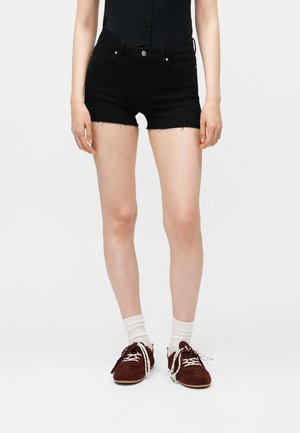 Donna con shorts di jeans neri a vita alta con orlo grezzo, calzini bianchi alla caviglia e sneakers bordeaux con lacci, in piedi su sfondo bianco.
