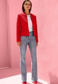 Roter, strukturierter Blazer, weißes Top, hellgraue ausgestellte Jeans und rote spitze High Heels. Gestylt mit Sonnenbrille und einfachem Schmuck vor einem rosa Hintergrund.
