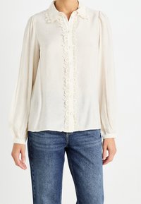 Blusa de crema claro con collar y borde de encaje, con botones en la parte delantera y mangas largas, combinada con jeans azules.