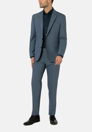 Hellblauem Anzug aus strukturiertem Stoff, einreihiger Blazer und schmal geschnittene Hose, kombiniert mit einem dunkelblauen Hemd und schwarzen Schuhen.