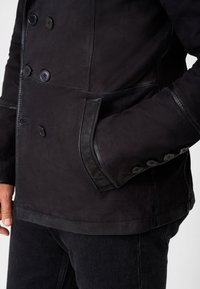Manteau en cuir noir avec un design à double boutonnage, de grands boutons, des poches latérales et une texture lisse, mate avec des coutures contrastées.