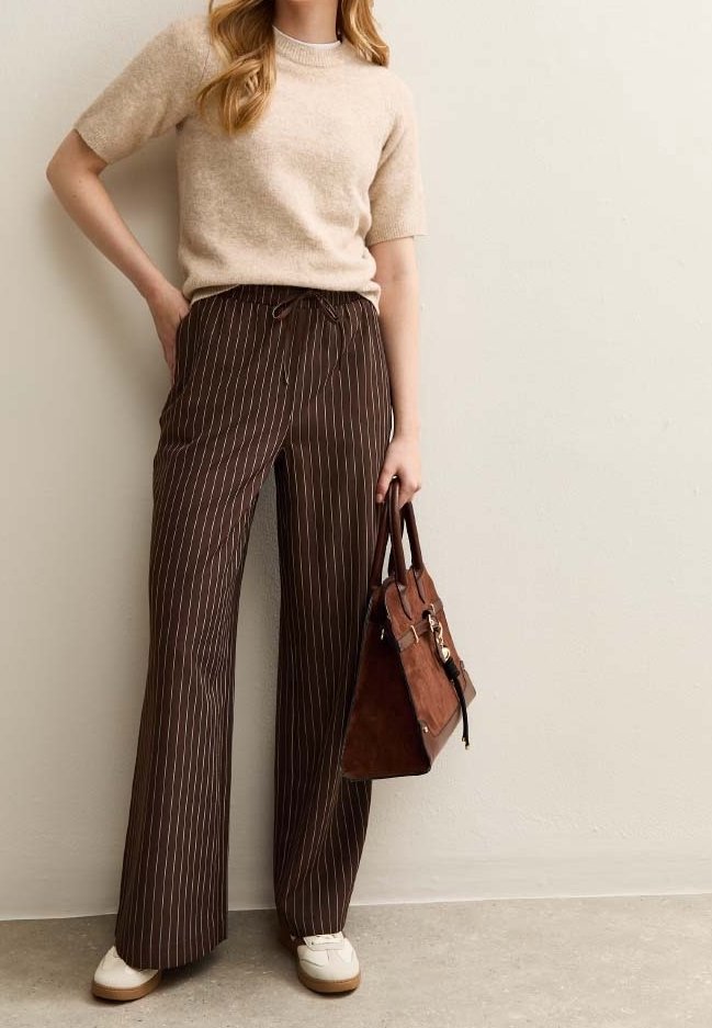 Femme portant un haut en maille beige à manches courtes, pantalon large marron à fines rayures, chaussures blanches, tenant un sac à main en cuir marron contre un mur uni.