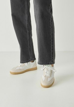 Baskets blanches en cuir et mesh, avec des accents beiges et une semelle en caoutchouc texturée, associées à un jean droit gris foncé.