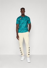 Turquoise shirt met korte mouwen met een patroon en een oranje logo, gecombineerd met crèmekleurige joggers met zwarte branding en witte sneakers.