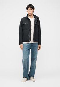 Νεαρός άντρας που φοράει μαύρο μπουφάν, μπεζ ζακέτα, φαρδιά μπλε τζιν παντελόνια και λευκά αθλητικά παπούτσια, στέκεται μπροστά σε απλό γκρι φόντο.