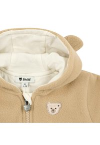 Lichtbruine fleece jacket met een capuchon, voorzien van een applicatie van een berengezicht, crèmekleurige voering en kleine berenoren op de capuchon. Voorzien van een rits aan de voorkant.