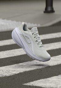 Γκρίζο αθλητικό παπούτσι με υφή στην επάνω πλευρά, λευκές λεπτομέρειες και ορατό σύστημα δέσιμος. Τοποθετημένο πάνω από ένα διάβαση πεζών σε γκρίζα επιφάνεια.