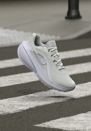 Grauer Sportschuh mit strukturiertem Obermaterial, weißen Akzenten und sichtbarem Schnürsystem. Über einem Fußgängerüberweg auf einer grauen Fläche platziert.