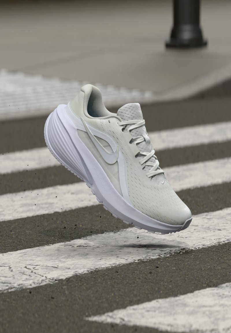 Γκρίζο αθλητικό παπούτσι με υφή στην επάνω πλευρά, λευκές λεπτομέρειες και ορατό σύστημα δέσιμος. Τοποθετημένο πάνω από ένα διάβαση πεζών σε γκρίζα επιφάνεια.