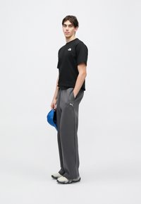 Μαύρο μπλουζάκι με λογότυπο, γκρίζο φόρμα με ελαστική μέση και εκρού παπούτσια. Κρατάει μπλε καπέλο, στέκεται μπροστά σε ανοιχτό φόντο.