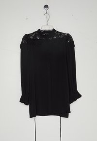 Blouse noire à manches longues avec détail en dentelle aux épaules, boutonnage devant, poignets froncés et col montant, suspendue sur un cintre transparent.