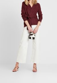 Femme portant un pull cache-cœur bordeaux et un pantalon large blanc, tenant un petit sac à main noir et blanc à motifs, chaussée de talons rouges à lanières.