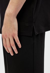 T-shirt preta de algodão com mangas curtas, fenda lateral e logo "ck" bordado na bainha. Mão repousada sobre o tecido exibe a textura.