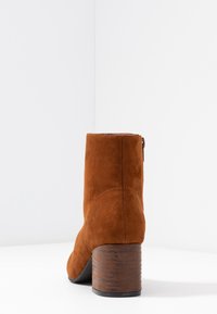 Bruine suède enkellaars met een gestructureerde blokhak van hout, ritssluiting aan de zijkant en een glad, afgerond teenontwerp.