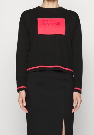 Haut noir à manches longues avec patch rectangulaire rouge portant l'inscription "ARMANI EXCHANGE" et bordure rouge aux poignets et à l'ourlet, associé à une jupe noire fendue à l'avant.
