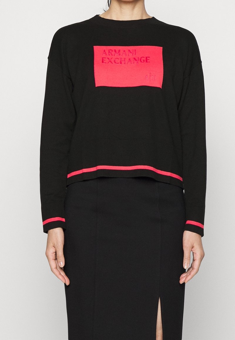 Haut noir à manches longues avec patch rectangulaire rouge portant l'inscription "ARMANI EXCHANGE" et bordure rouge aux poignets et à l'ourlet, associé à une jupe noire fendue à l'avant.