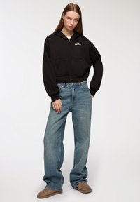 Felpa nera corta con zip e tasche frontali, abbinata a jeans leggeri a vita alta di colore azzurro. Il modello indossa scarpe slip-on beige.