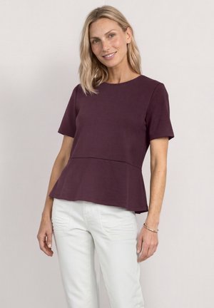 Femme aux cheveux blonds mi-longs portant un top péplum violet foncé à manches courtes et un pantalon blanc, souriant légèrement devant un fond uni.