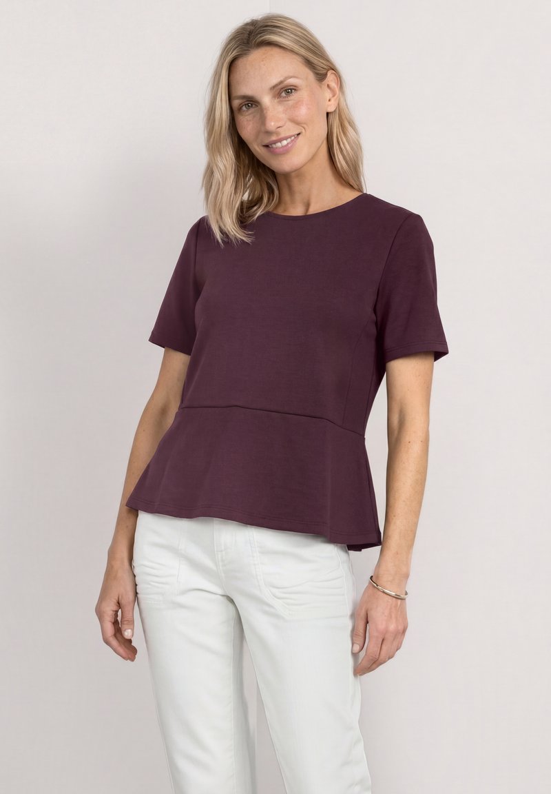 Femme aux cheveux blonds mi-longs portant un top péplum violet foncé à manches courtes et un pantalon blanc, souriant légèrement devant un fond uni.