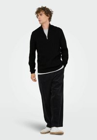 Junger Mann in einem schwarzen Strick-Zip-Pullover, lockeren schwarzen Hosen und weißen Sneakers, der mit einer Hand in der Tasche steht.