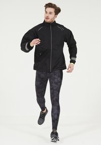 Veste coupe-vent noire avec des accents réfléchissants, associée à des leggings en camouflage sombre et des chaussures de sport noires, dans une pose dynamique.