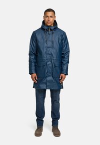 Marineblauwe waterdichte jas met een capuchon, twee voorkruiken, verstelbare trekkoorden en een gladde, glanzende afwerking; gedragen over een blauwe spijkerbroek.
