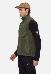 Groene fleece vest met hoge kraag en ritssluiting, voorzien van een borstzak en zijzakken met rits. Draagt over een zwart shirt met lange mouwen.