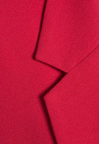 Punainen kangas, jossa on teksturoitu pinta, joka koostuu päällekkäisistä osista ja kulmikkaista reunoista, mikä antaa sille sileän, kreppimäisen ilmeen.