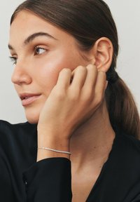 Fiatal barna hajú nő lófarokkal, fekete felsőt és finom ezüst karkötőt visel, kezét a füle közelében tartja pihenés közben.