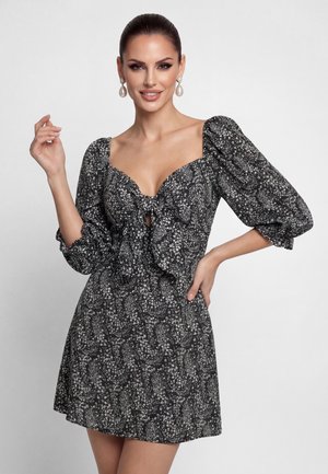 Femme portant une robe à fleurs noire et blanche avec des manches bouffantes et un nœud à l'avant, posant avec une main sur la hanche et l'autre levée.