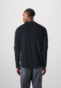 Marineblå hettegenser med lange ermer, ribbestrikkede mansjetter og nederkant, samt et V-formet stikklag bak. Kombinert med grå denimjeans.
