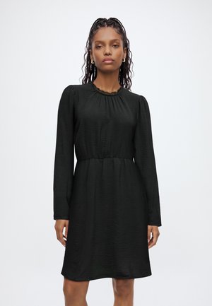 Femme portant une robe noire à manches longues avec une taille froncée et un col rond bordé de dentelle, debout devant un fond blanc uni.