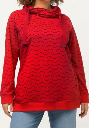 Frau trägt einen roten Kapuzenpullover mit schwarzen wellenförmigen Streifen und Kordeln, kombiniert mit hellblauen Jeans vor einem einfarbigen Hintergrund.