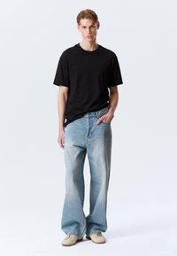 T-shirt noir à col rond, coupe décontractée. Jean large bleu clair avec des zones décolorées. Baskets beiges avec un détail de lacets. Design minimaliste.