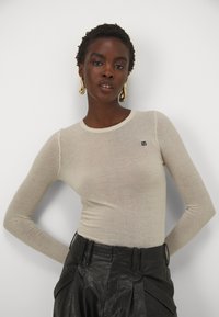 Lys beige langermet topp laget av et gjennomsiktig materiale med rund hals; har et lite logodesign på brystet. Kombinert med svarte skinnbukser.