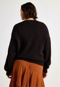 Žena s krátkými tmavými vlasy, oblečená v volném černém svetru a sukni v cihlové barvě se výrazným volánkovým detailem, zobrazená zezadu.