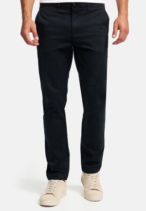 Uomo che indossa pantaloni neri a taglio dritto e sneaker beige con lacci, in piedi contro uno sfondo bianco uniforme.