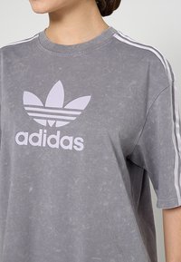 Camiseta de algodón gris oversize con acabado texturizado, logo de Adidas en blanco y detalles de tres rayas blancas en los hombros.
