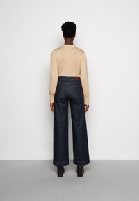 Person som står med ryggen vänd mot kameran, iklädd en beige långärmad topp, högmidjade vida mörka jeans och svarta klackskor mot en vit vägg.