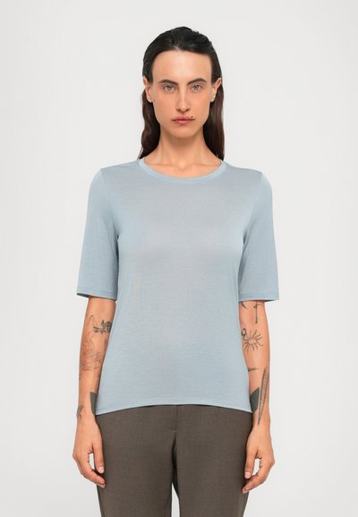 Persoon met lang donker haar, gekleed in een lichtblauw shirt met korte mouwen en een broek in bruin, met tatoeages op beide onderarmen tegen een effen achtergrond.