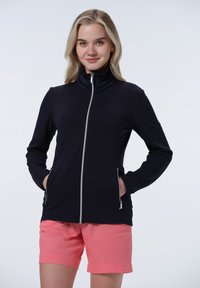 Veste zippée bleu marine avec un design texturé, col montant et deux poches avant, associée à un short rose clair. Fond épuré.
